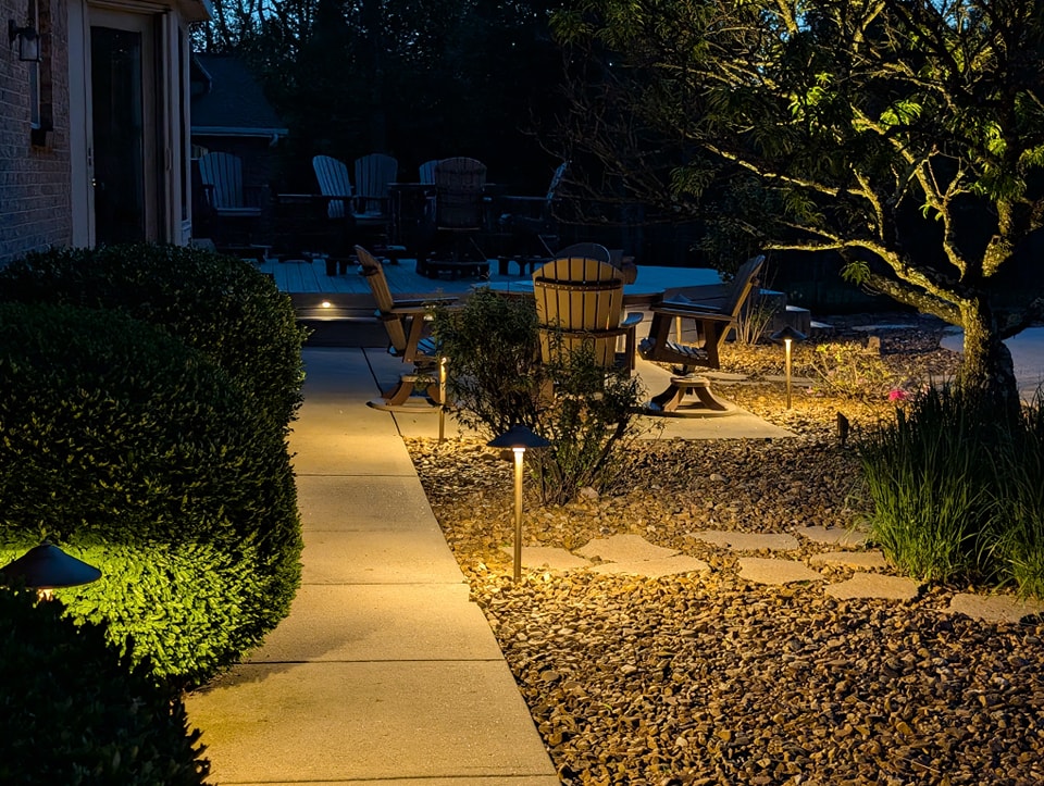 Pathway illuminated by stylish outdoor lighting, surrounded by landscaped bushes and rocks, leading to a cozy seating area with wooden chairs.