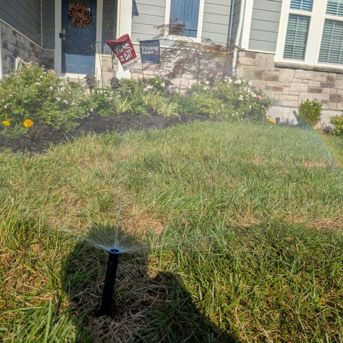 Sprinkler system in operation, watering green lawn with flowers and house in background, illustrating irrigation maintenance services in Northern Kentucky.